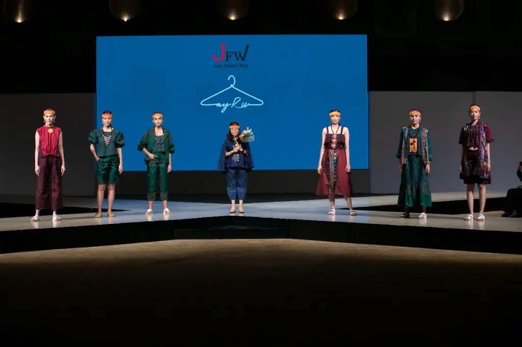 Zayru showcase at Jogja Fashion Week — designer takes the stage between eight models in earthy red and green aksara batik, JFW × Zayru logo lit on the LED backdrop.